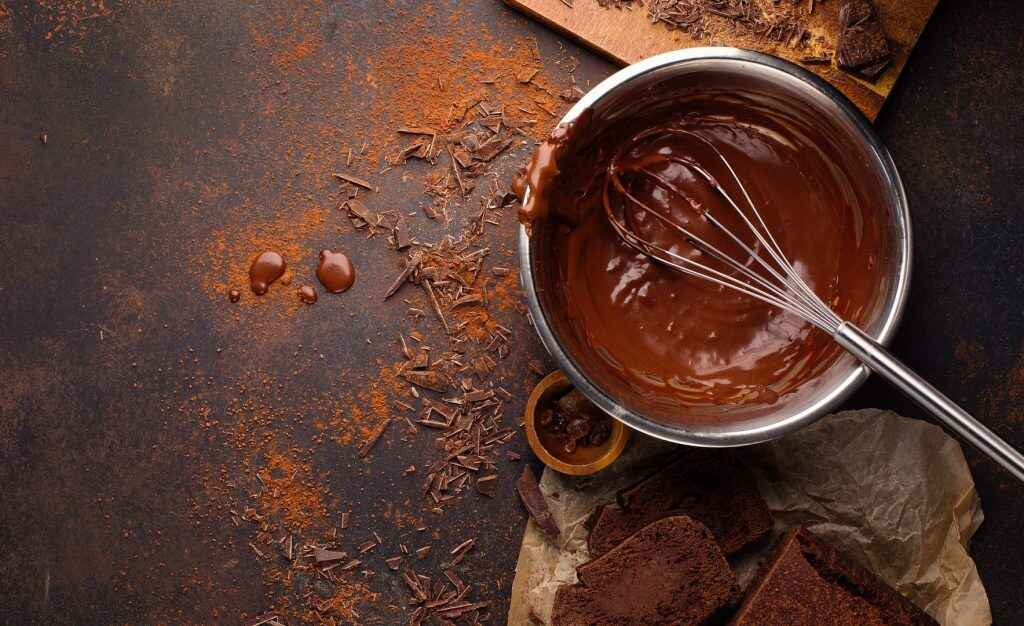 A Baking bowl filled with melted chocolate and a whisk. Surrounded by chocolate drops and chocolate cake, showing everything you need to know about chocolate cake.